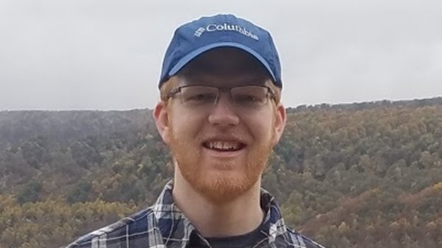 portrait of Nate Kratzer standing outside in front of a hill covered in trees